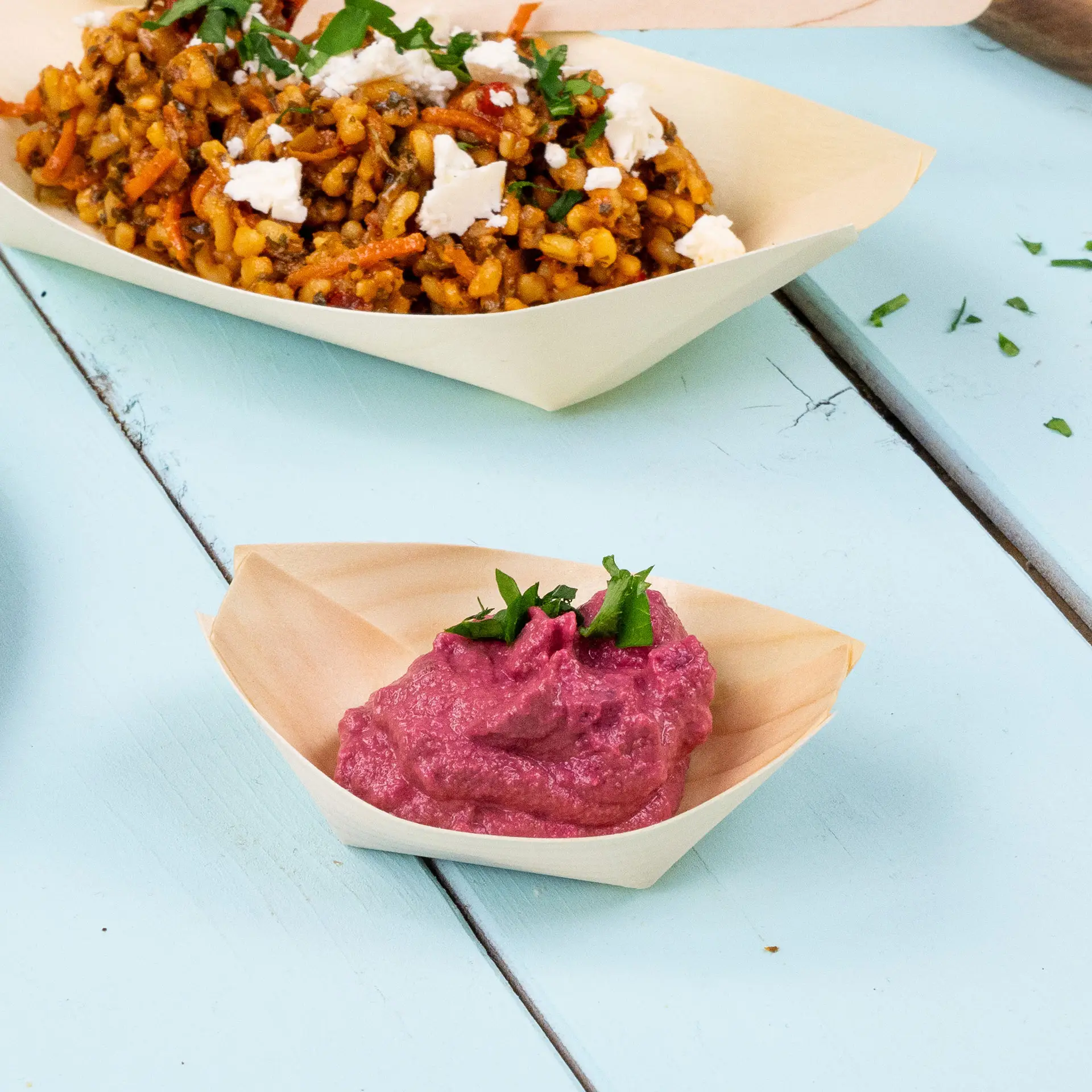 "Boat-shaped wooden bowl holding pink dip, garnished with fresh herbs; background features a bowl of mixed dish."
